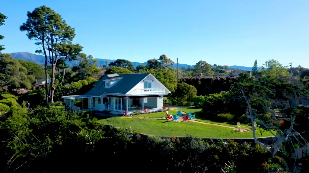 A white house with a dark roof on a grassy bluff overlooking mountains.
