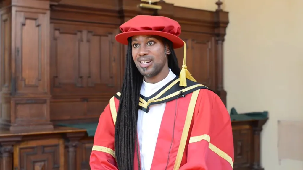 Jason Arday in academic regalia, wearing a red hat and gown with gold trim, standing in front of wooden paneling.