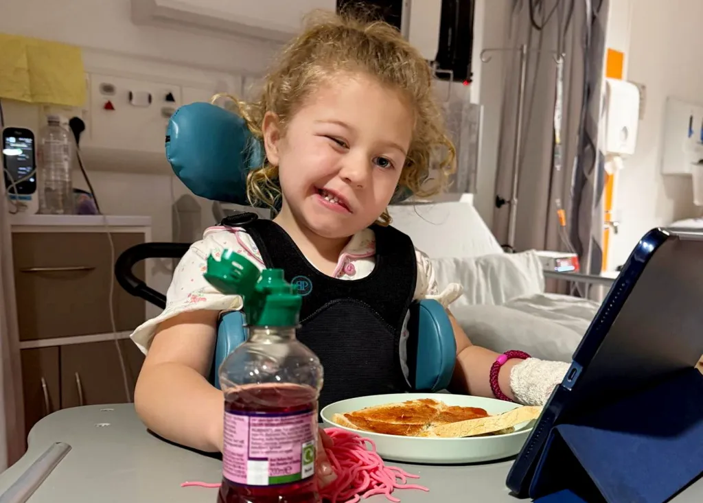 Olivia Elliot, a four-year-old girl, wearing a back support, sits at a hospital table with toast and a drink, winking at the camera.