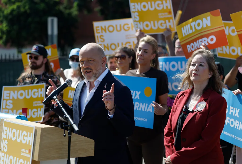 California Republican gubernatorial candidate Steve Hilton speaks at a press conference next to fellow politician Gloria Romero, with supporters behind them holding campaign signs.