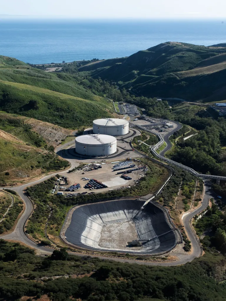 Aerial view of Sable Offshore's Las Flores Canyon facility with storage tanks, a basin, and winding roads in hilly terrain near the ocean.
