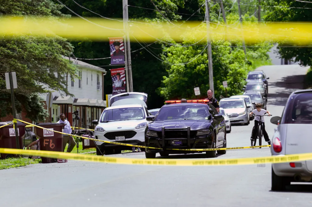 Police cars and yellow crime scene tape block off a street with homes and trees in the background.