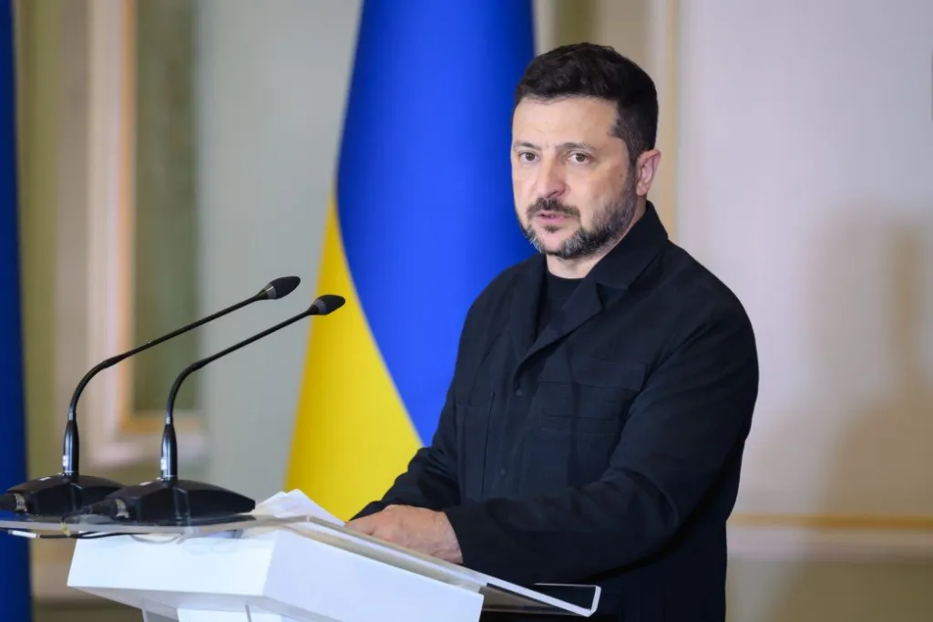 Ukrainian President Volodymyr Zelenskyy speaking at a podium with microphones, with the Ukrainian flag in the background.