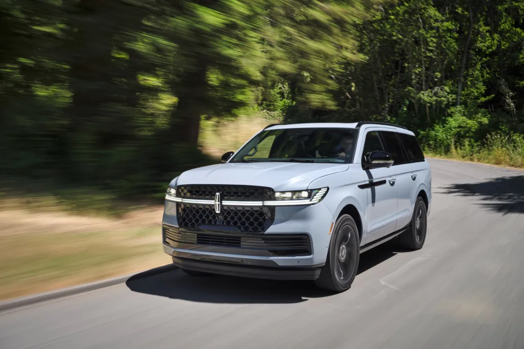 The 2026 Lincoln Navigator, a light gray luxury SUV, drives along a road with blurred green trees in the background.