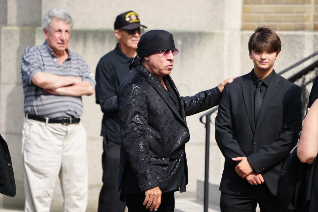 Stevie Van Zandt, in a black bandana and patterned black jacket, places a hand on the shoulder of a young man in a black suit.