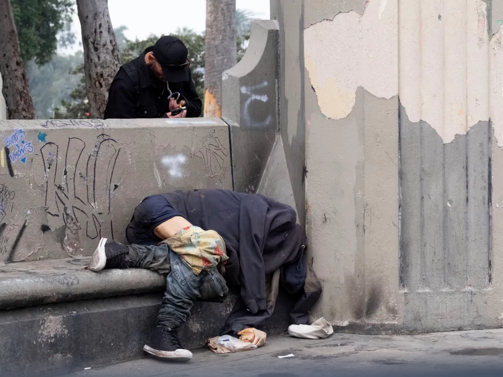 A man lights a crack pipe as another person lies on the ground next to a wall covered in graffiti.