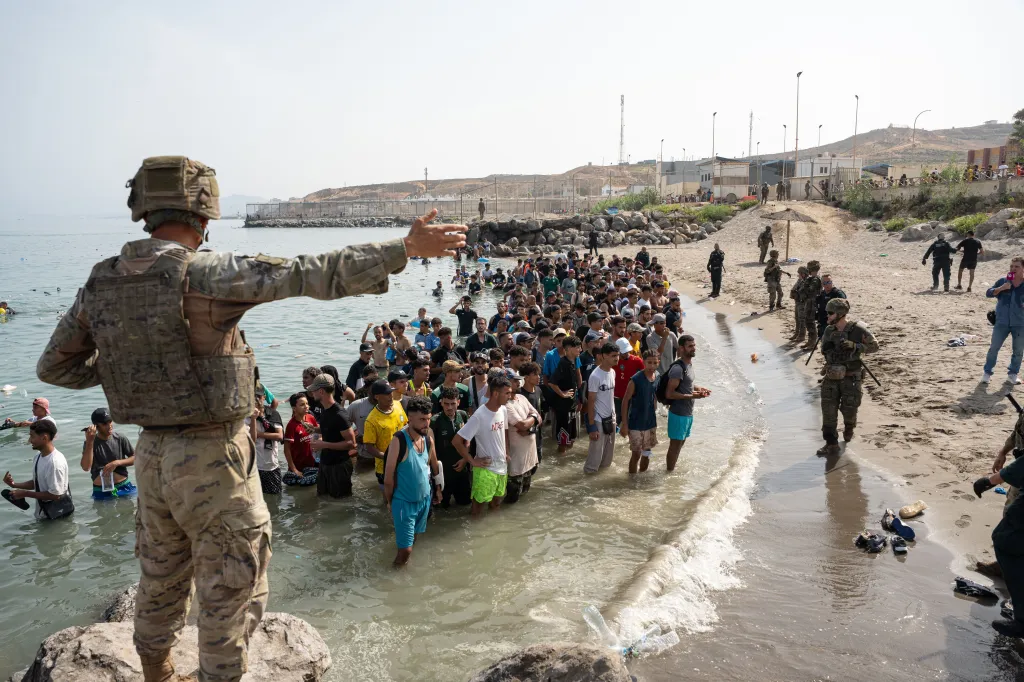 Migrants standing in the water at the Ceuta border with a soldier in camouflage pointing towards them.