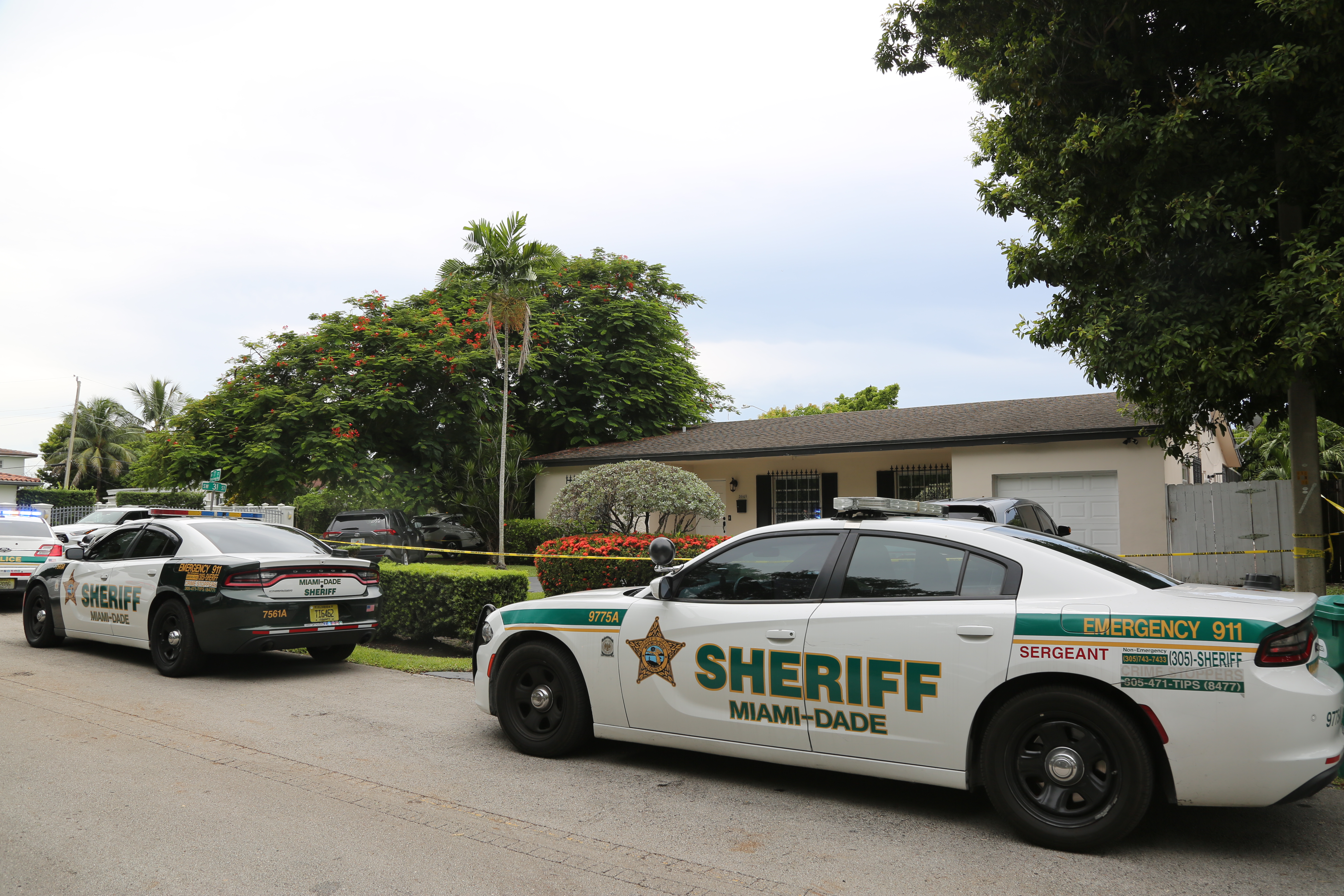 Two Miami-Dade Sheriff's cars parked outside a house with yellow crime scene tape across the lawn.