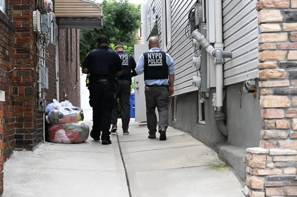 Three NYPD officers walk down an alleyway next to a building with siding and exposed pipes.