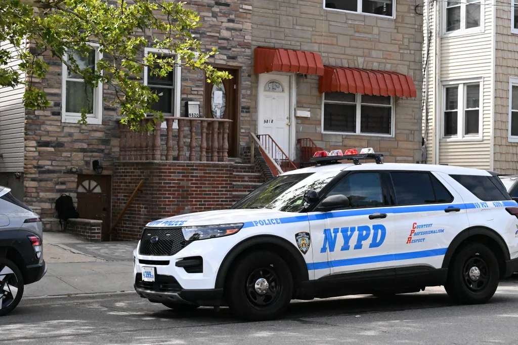 A white NYPD police vehicle with red and blue lights sits parked on a street in front of a residential building.