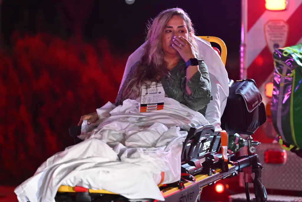 A woman bundled in a white blanket sits on a stretcher at night, her hand covering her mouth as if distraught or crying, with the red light of an emergency vehicle in the background.