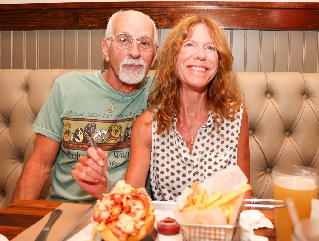 A man and a woman sitting at a restaurant with lobster rolls, fries, and drinks.