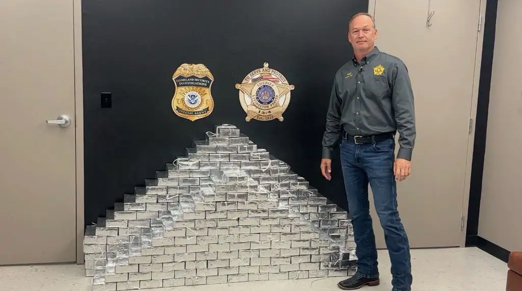 A uniformed officer stands next to a large stack of drug bundles.