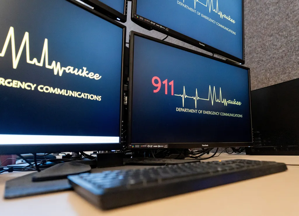 A 911 call station with multiple monitors displaying "911" and "Department of Emergency Communications" with an ECG line graphic.