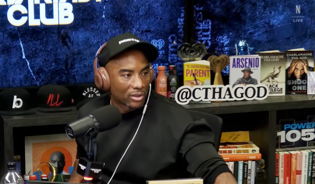 Charlamagne tha God in a black cap and shirt, wearing headphones and speaking into a microphone.