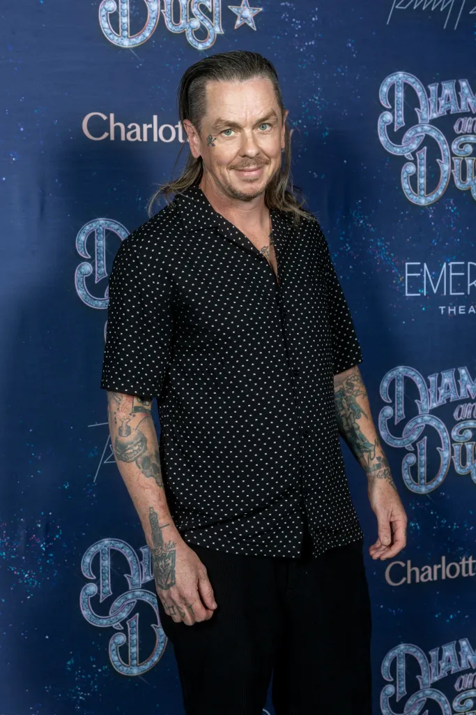 Joel Madden poses in a black patterned shirt with tattoos on his arms and a skull and crossbones tattoo near his eye.