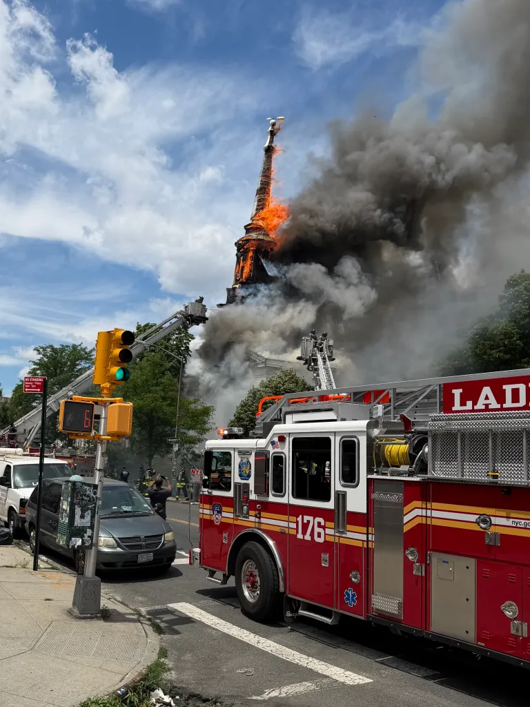 Firefighters extinguishing a fire in the steeple of a building with a fire truck in the foreground.