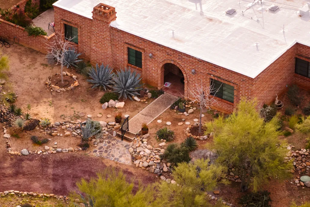 An aerial view of a brick house with a flat white roof and a desert-like garden with rocks, cacti, and arid plants.