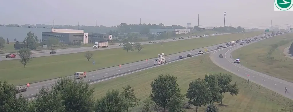 Highway traffic with a work vehicle, several orange cones, and a police car with flashing lights.