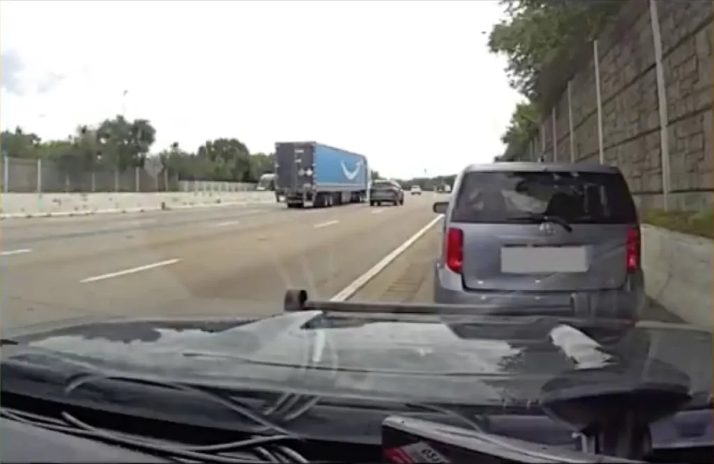 Dashcam view of a gray car following another gray car closely on a highway, with an Amazon semi-truck further ahead.