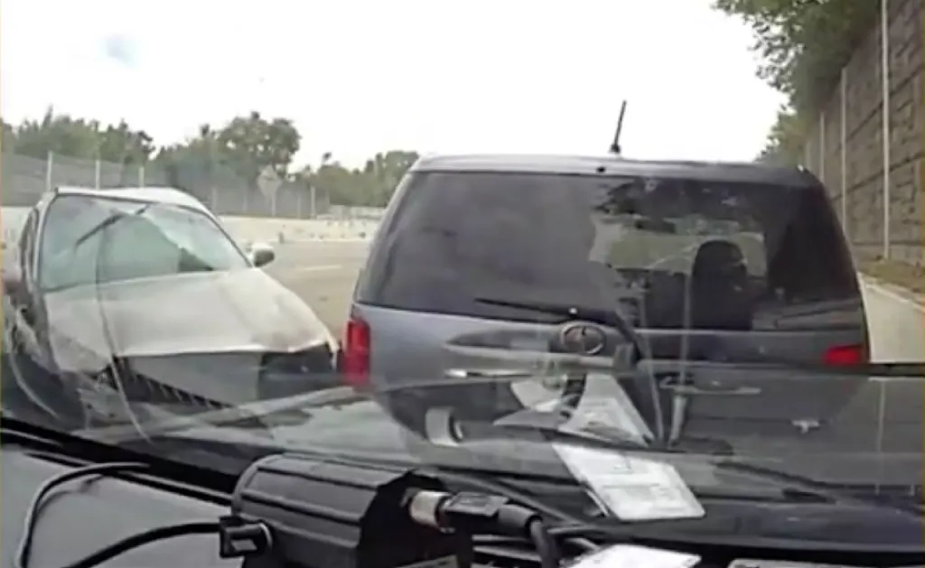 A damaged car with a crumpled hood stopped behind an SUV on a highway, with trees and a sound wall in the background.