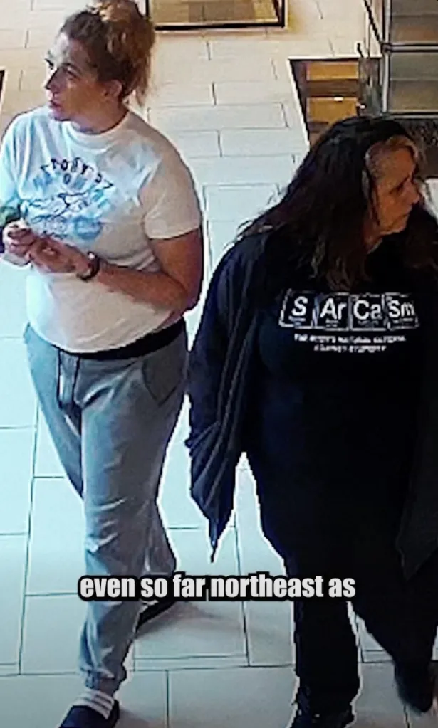 Two women walking, one in a white shirt and gray pants, the other in a black long-sleeved shirt with the word "Sarcasm" on it.