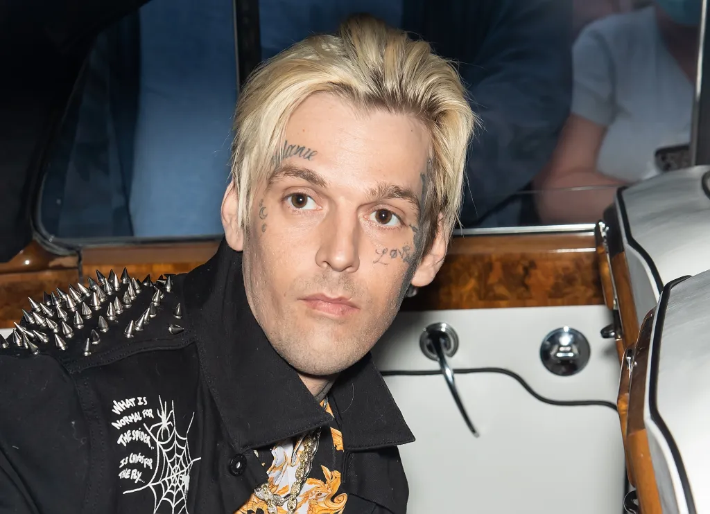 Aaron Carter, with bleach blonde hair and face tattoos, wearing a black studded jacket and patterned shirt, looks directly ahead.
