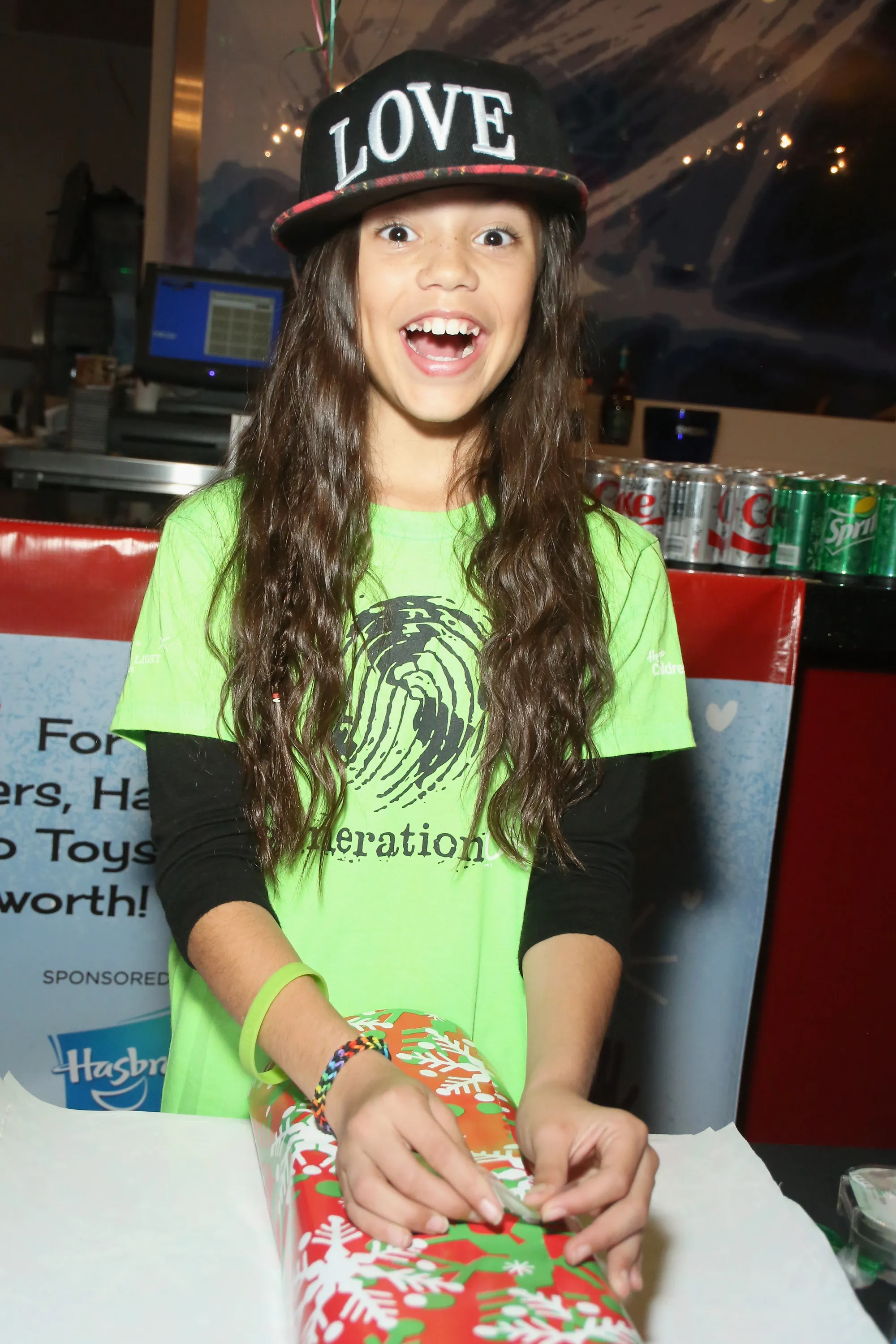 Actress Jenna Ortega in a green shirt and "LOVE" hat, enthusiastically wrapping a present.