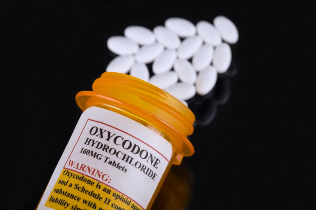 Prescription bottle of Oxycodone Hydrochloride 160MG tablets with a warning label on a black surface next to loose white pills.