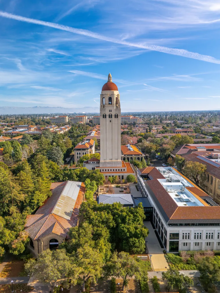 Aerial view of Stanford University in Palo Alto, California, with the Hoover Tower and campus buildings surrounded by trees under a blue sky.
