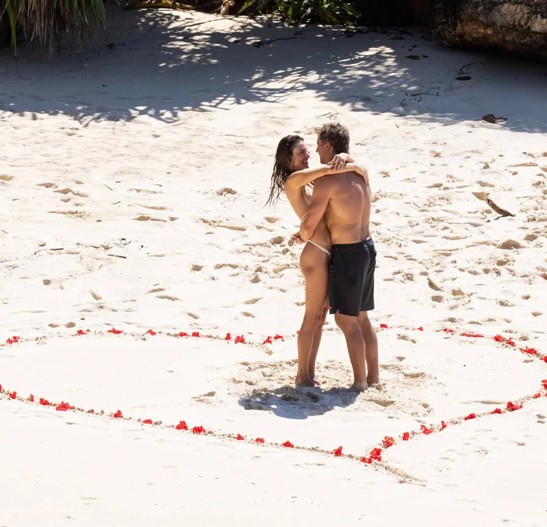 Alessandra Ambrosio and Buck Palmer embracing on a sandy beach inside a heart outline made of red flower petals.