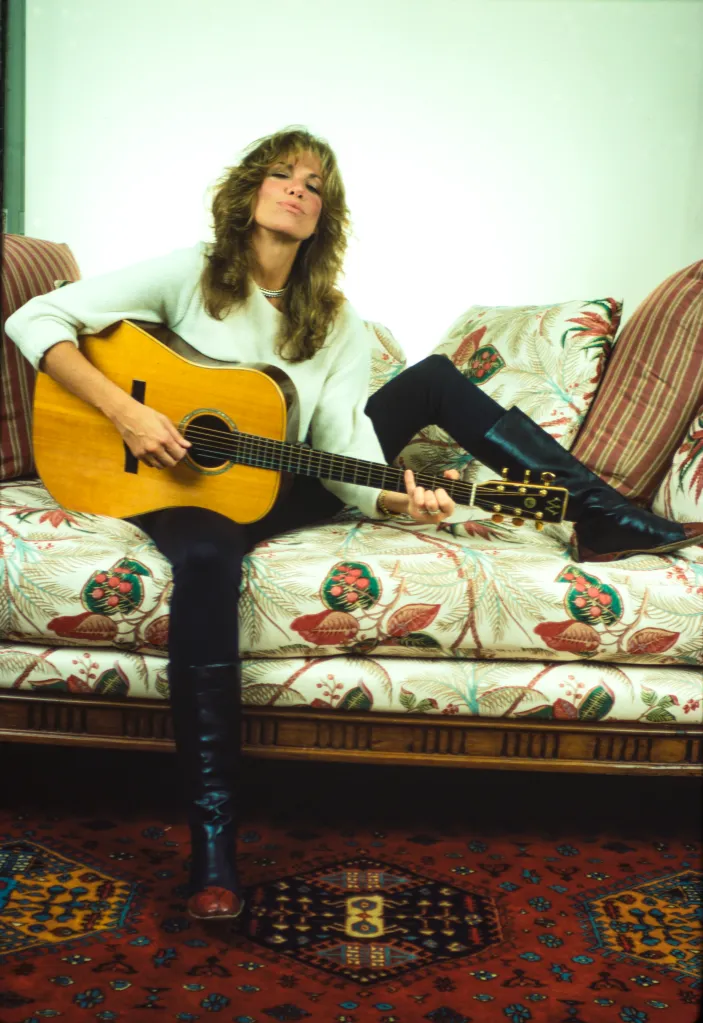 Carly Simon sitting on a floral sofa with a guitar, wearing a white sweater, black pants, and black boots.