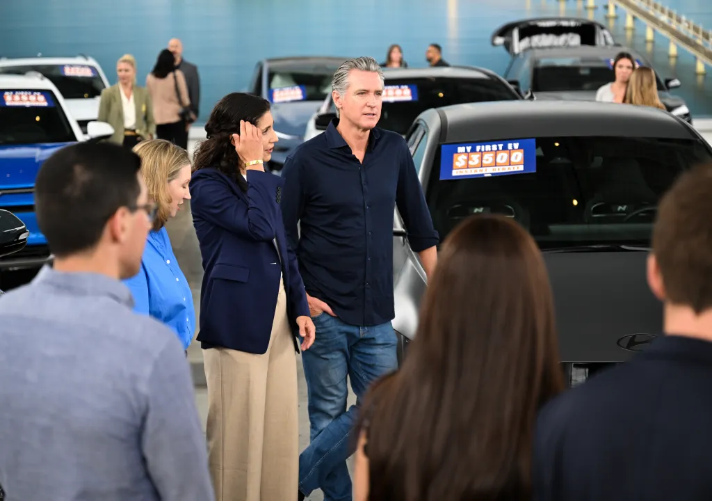 Governor Gavin Newsom speaking at a press conference with several electric vehicles, one showing a $3500 instant rebate.
