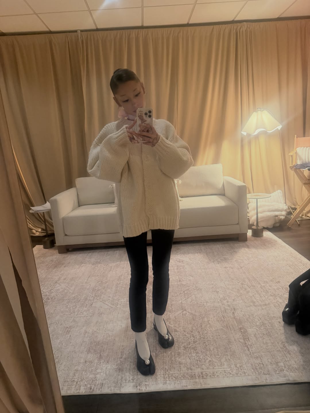 Ariana Grande taking a mirror selfie wearing a cream cardigan, black pants, white socks, and black slingback flats.