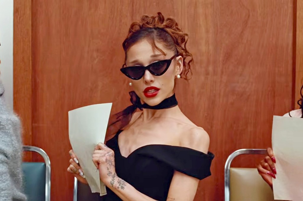 Ariana Grande in a black off-the-shoulder dress, cat-eye sunglasses, and a black choker, holding papers.
