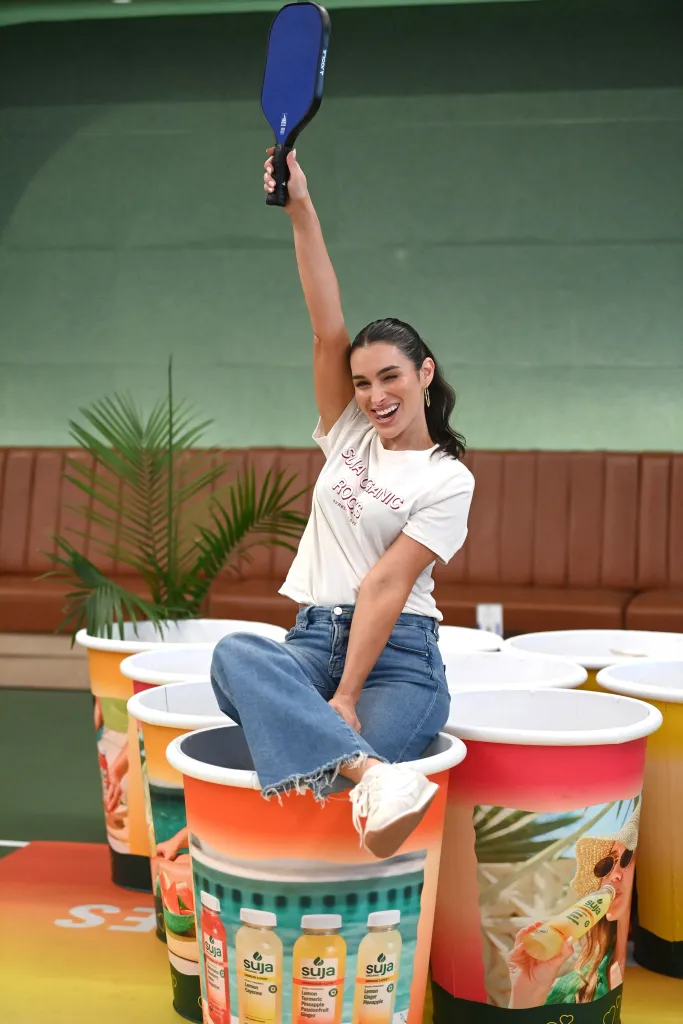 Ashley Iaconetti holds a pickleball paddle overhead while sitting in an oversized Suja Organic cup.