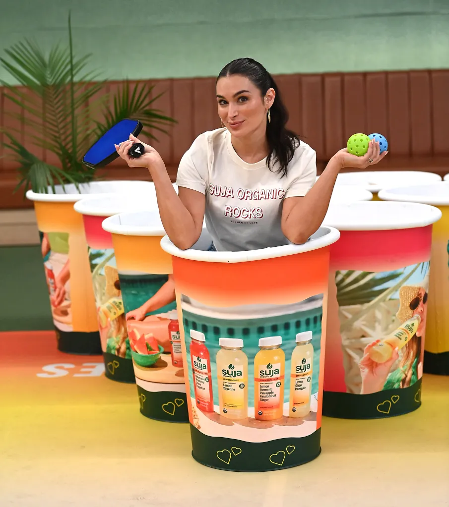 Ashley Iaconetti posing inside a large Suja Organic cup, holding a pickleball paddle and two pickleballs.