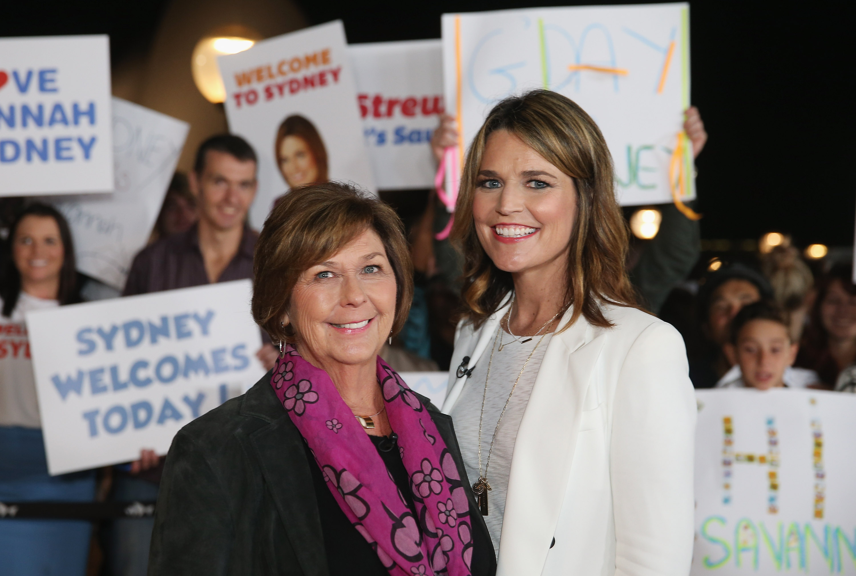 Savannah Guthrie and her mother Nancy Guthrie smiling at the camera with a crowd holding signs in the background.