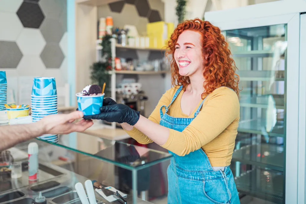 A smiling redhead woman in a denim apron and yellow shirt serving ice cream in a blue cup to a customer.
