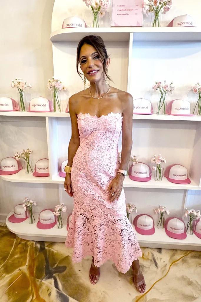 Bethenny Frankel posing in a pink lace dress with "supermodel" branded hats and small flower arrangements displayed on shelves behind her.
