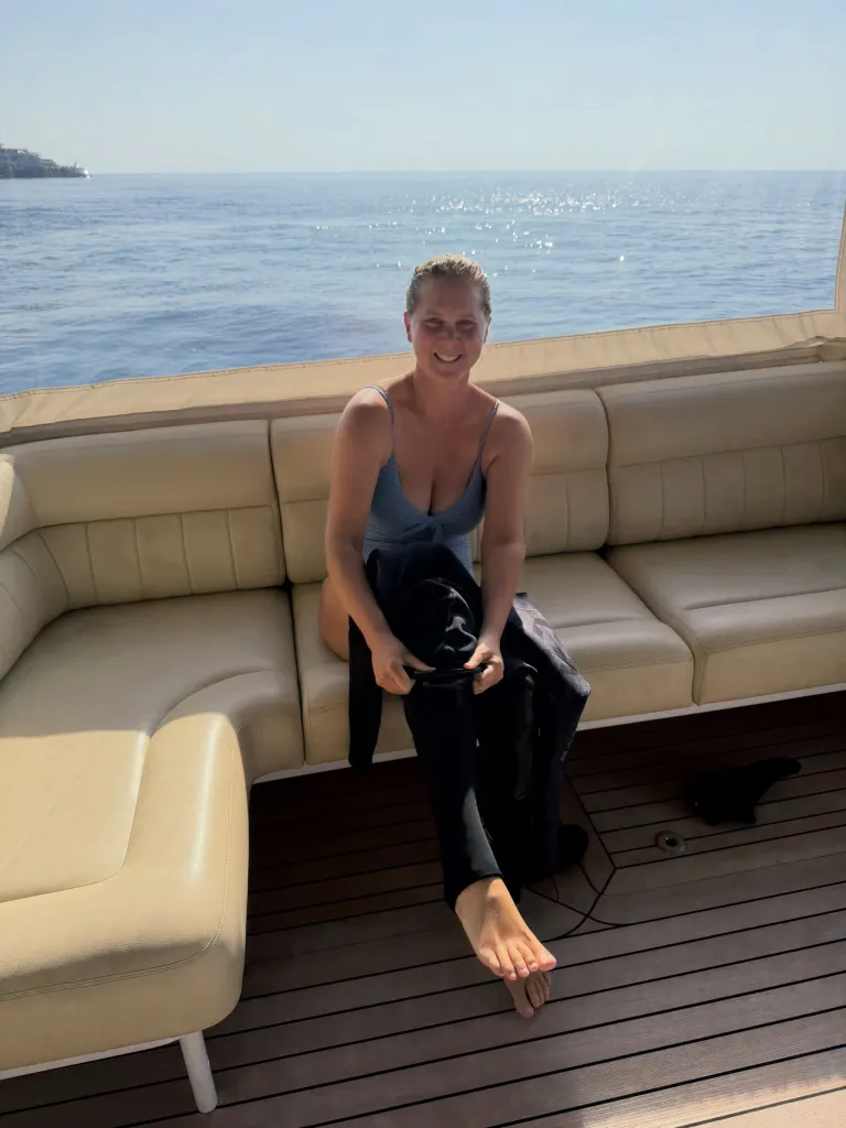 Amy Schumer in a light blue swimsuit on a boat, holding a black wetsuit over her lap.