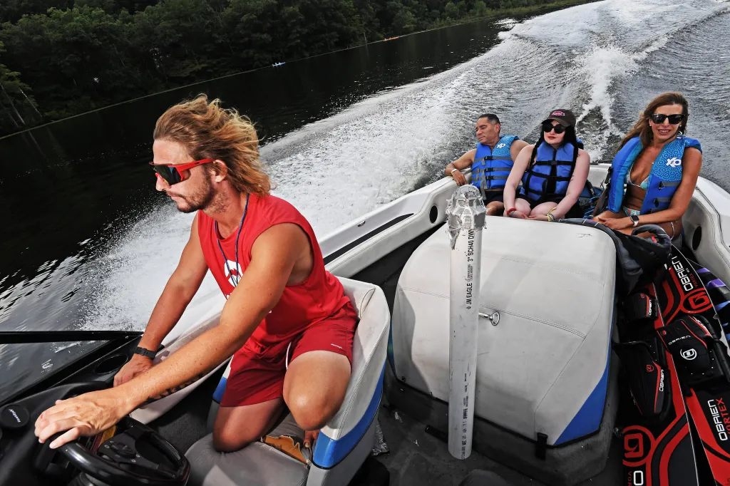 A man drives a motorboat while three passengers in life vests sit behind him, creating a wake on the lake.