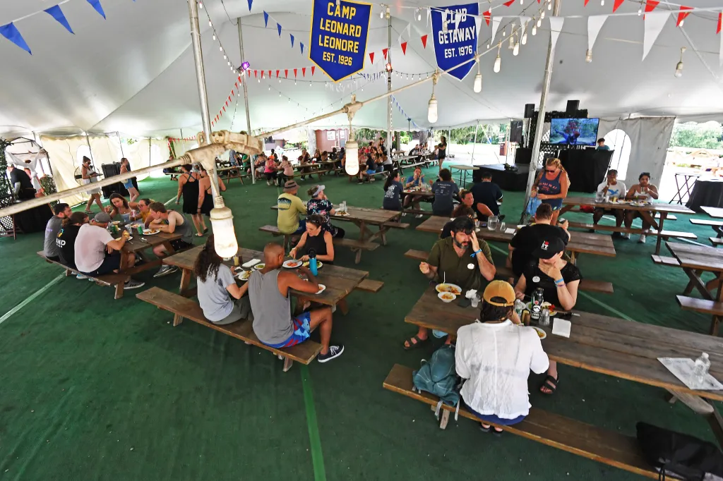 People eating and socializing at picnic tables under a large tent with banners for "Camp Leonard Leonore" and "Club Getaway."