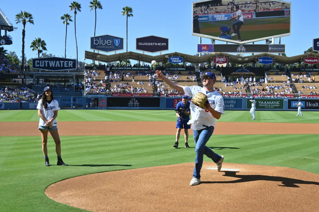 A man in a Dodgers jersey throws a baseball from the pitcher's mound while a woman stands nearby, with palm trees and stadium seating in the background.
