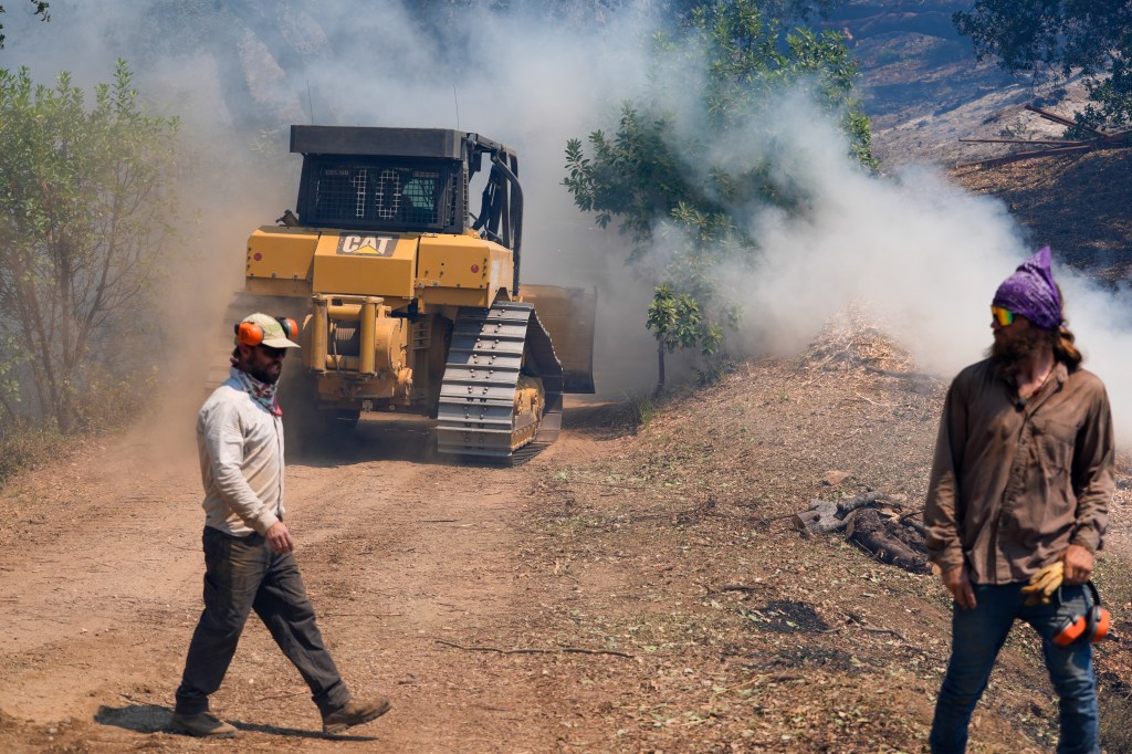 A bulldozer clearing land in a smoky area with two men standing nearby during the Timber Fire in Big Sur, California.