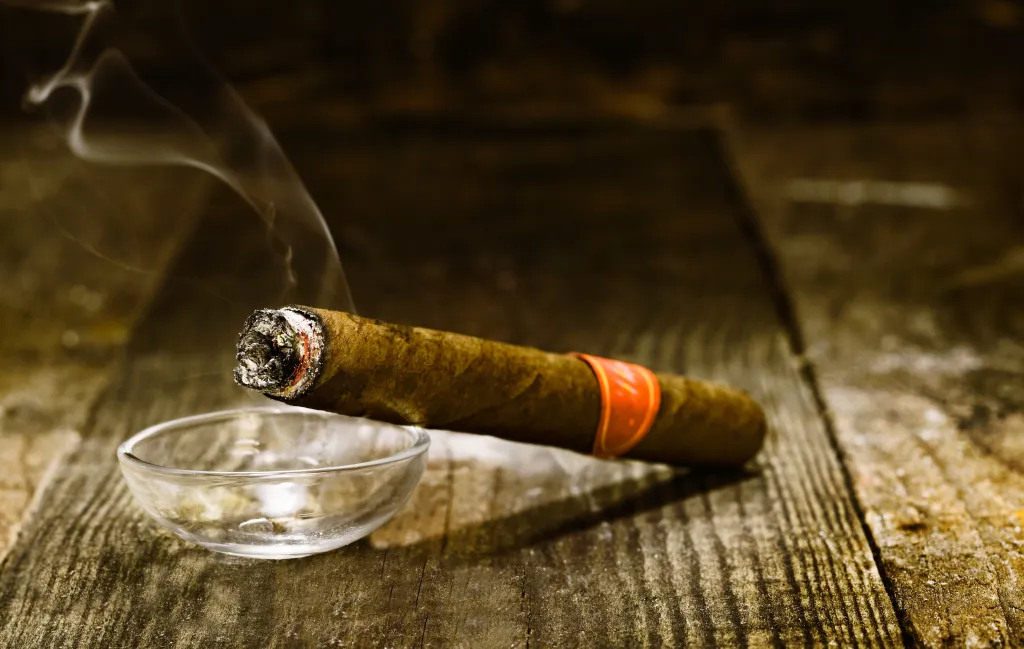 Burning handmade luxury Cuban cigar resting on an ashtray on an old wooden countertop.