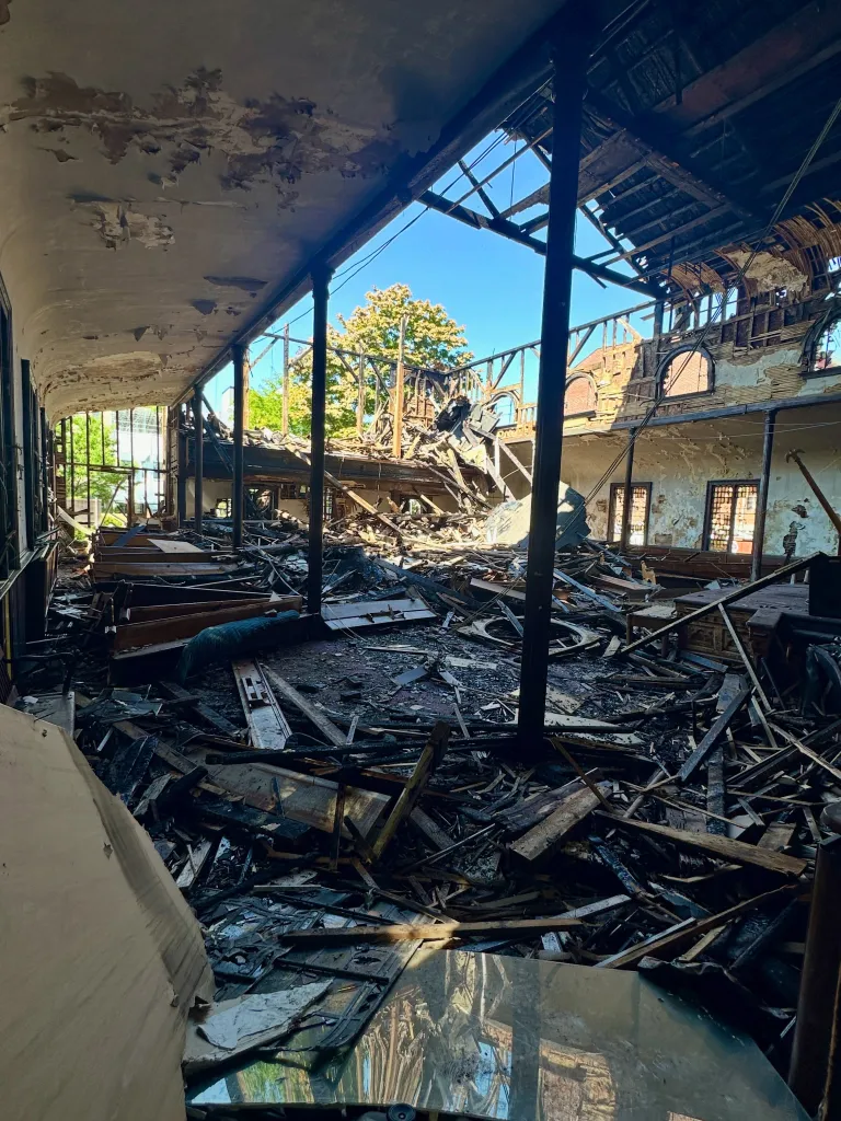 Interior of a severely damaged and collapsed building with debris on the floor and a visible blue sky through the destroyed roof.