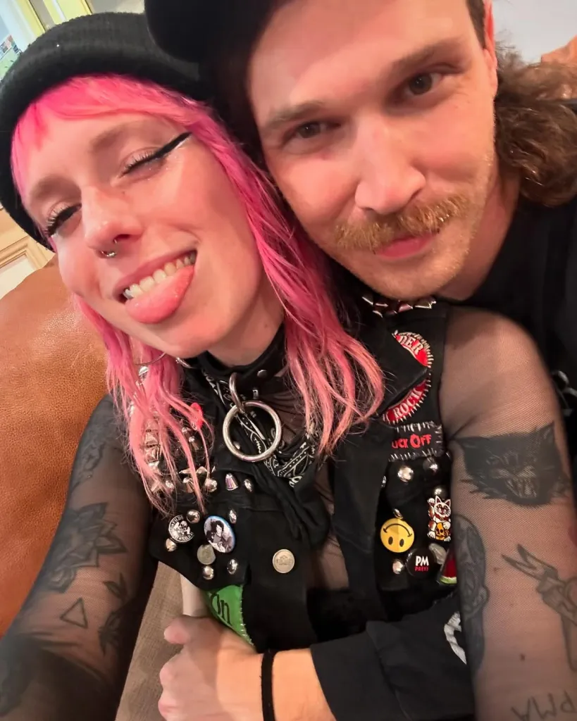 A woman with pink hair and a man with a mustache pose together.