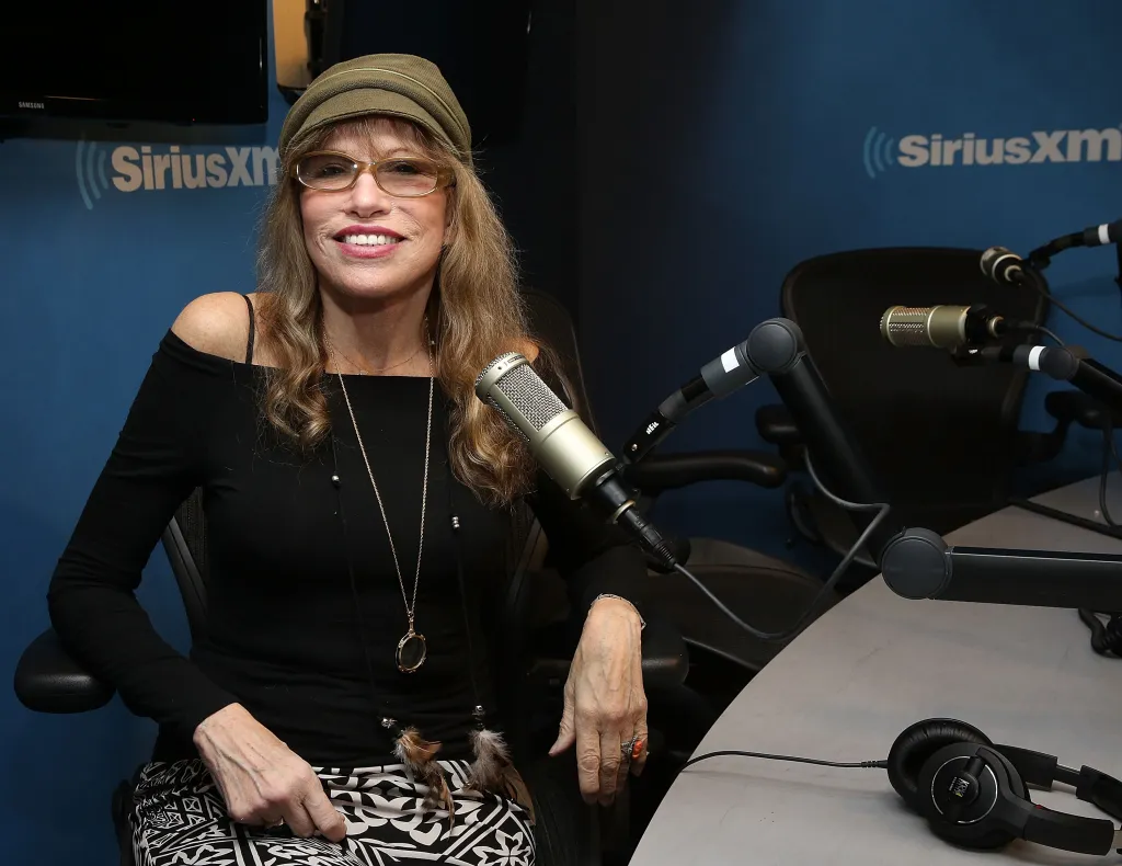 Carly Simon in the SiriusXM studio, seated and smiling, wearing a khaki hat, glasses, and a black off-the-shoulder top.