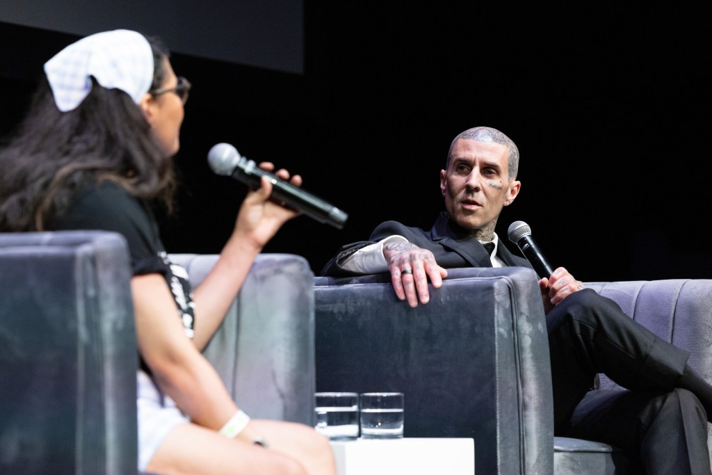 Travis Barker sitting in a chair holding a microphone and looking at an interviewer.